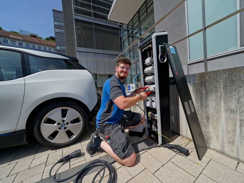Ein Techniker kniet vor einer geöffneten Ladestation neben einem Elektroauto und arbeitet daran.