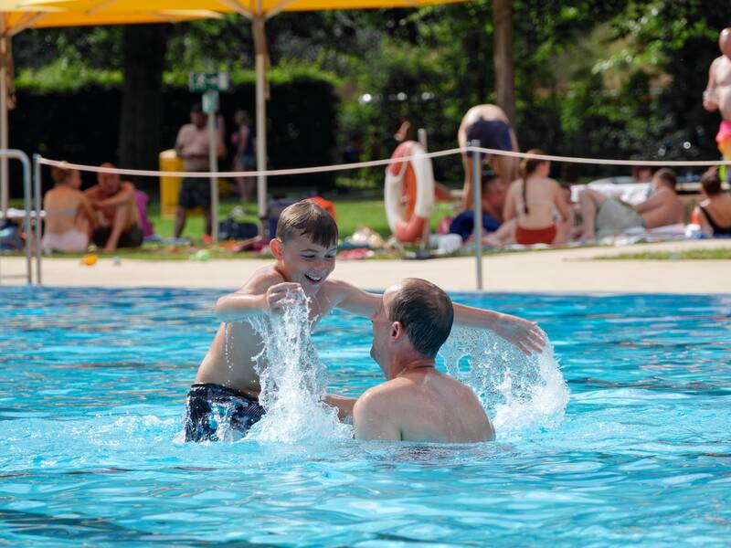 Ein Junge spielt fröhlich mit einem Erwachsenen im Wasser eines Schwimmbeckens, während im Hintergrund andere Menschen sich am Beckenrand aufhalten.