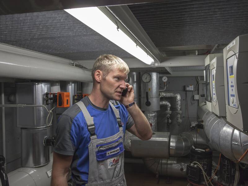 Ein Mann in Arbeitskleidung telefoniert in einem Technikraum mit Rohren und Kontrollgeräten.