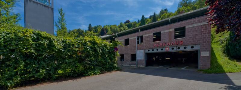 Ein zweistöckiges Parkhaus aus rotem Backstein mit der Aufschrift "PARKHAUS" befindet sich neben einer grünen Hecke und einem Turm vor einem bewaldeten Hügel unter blauem Himmel.