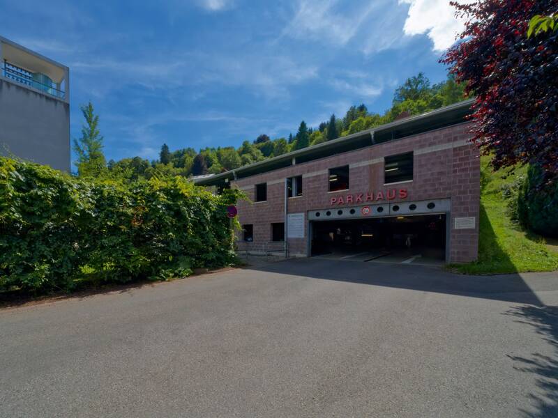 Ein zweistöckiges Parkhaus aus rotem Backstein mit der Aufschrift "PARKHAUS" befindet sich neben einer grünen Hecke und einem Turm vor einem bewaldeten Hügel unter blauem Himmel.