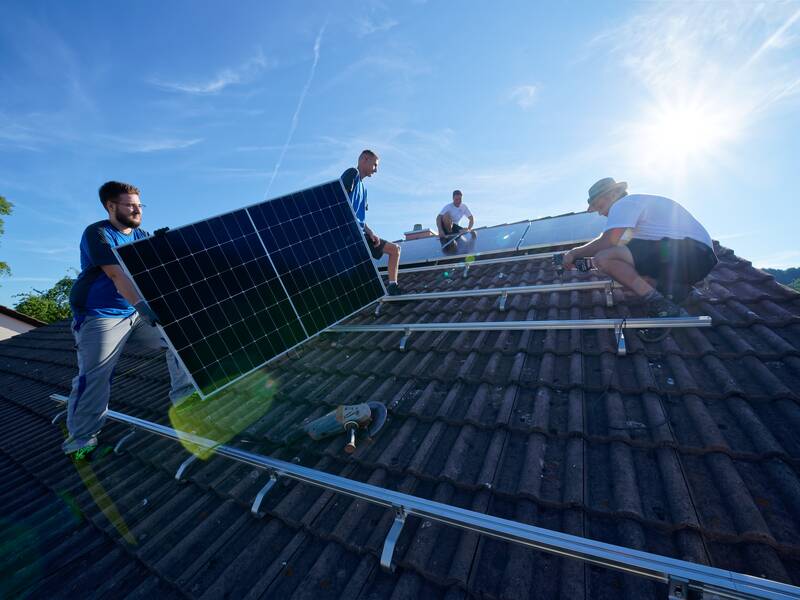 Vier Personen installieren Solarmodule auf einem Hausdach bei sonnigem Wetter.
