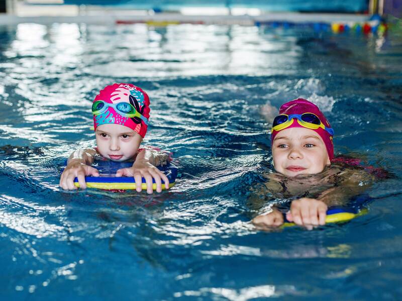 Zwei Kinder mit Schwimmbrillen und -kappen sind in einem Schwimmbecken und halten Schwimmbretter.