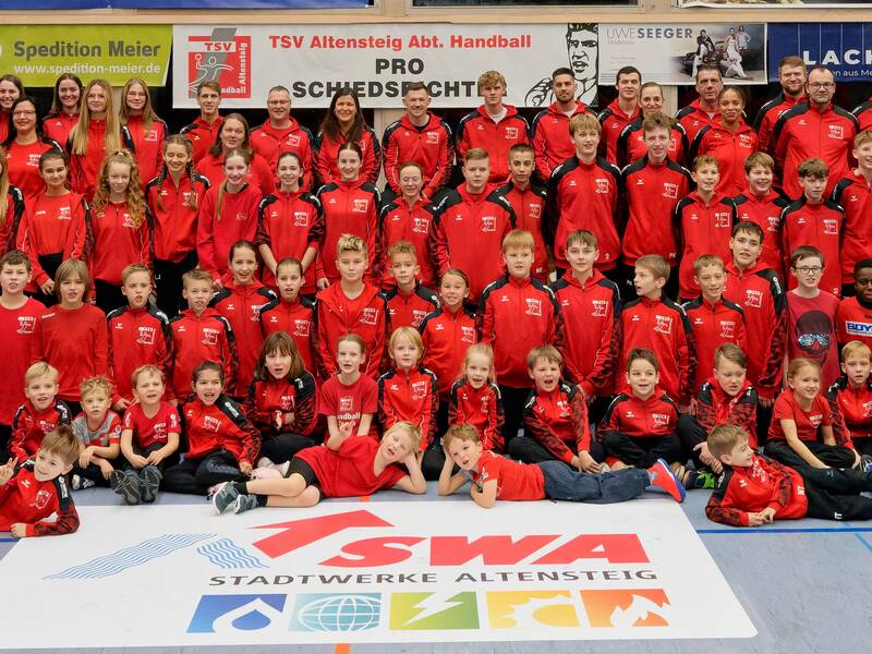 Eine große Gruppe von Menschen in roten Sportanzügen posiert lächelnd in einer Sporthalle für ein Gruppenfoto.