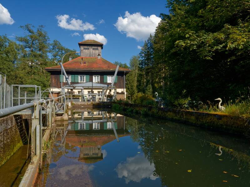 Ein Wasserkraftwerk mit rotem Dach und Holzverkleidung spiegelt sich in einem schmalen Kanal, der von üppigem Grün umgeben ist, wider.