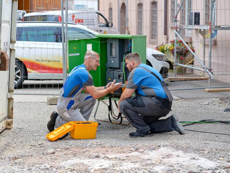 Zwei Männer in Arbeitskleidung arbeiten an einem grünen Verteilerkasten auf einer Baustelle.
