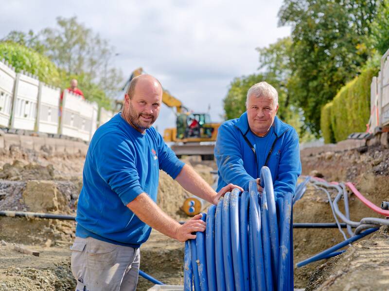 Zwei Männer in blauen Arbeitsjacken stehen in einem Graben und halten ein Bündel blauer Rohre, während im Hintergrund eine Baustelle sichtbar ist.