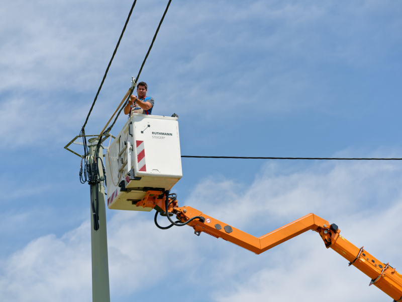 Ein Mann steht in einem Arbeitskorb eines orangefarbenen Hebearms und arbeitet an einer Leitung auf einem Strommast vor blauem Himmel.