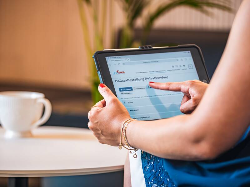 Eine Person bedient ein Tablet, auf dem eine Webseite zur Online-Bestellung für Privatkunden geöffnet ist, während im Hintergrund eine Tasse auf einem Tisch steht.