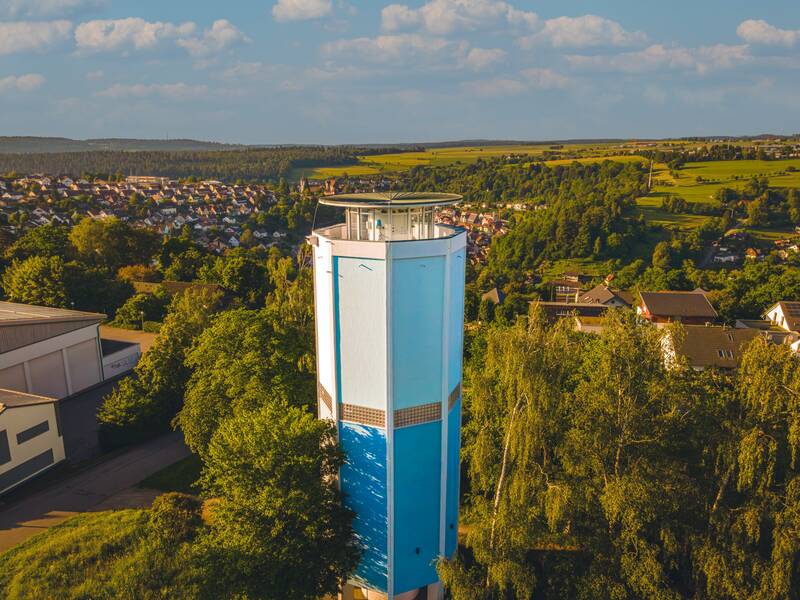 Ein blauer Wasserturm erhebt sich über eine grüne Landschaft mit einer Stadt im Hintergrund.