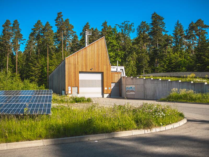 Ein modernes, holzverkleidetes Gebäude mit einem begrünten Dach steht inmitten einer Waldlandschaft neben einer kleinen Solaranlage.