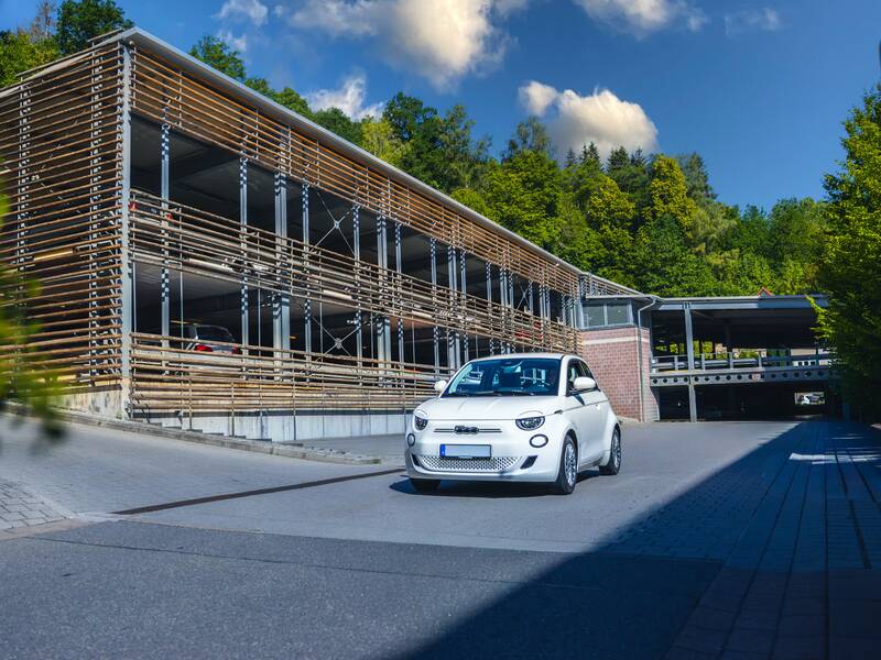Ein weißes Auto fährt auf einer Straße an einem mehrstöckigen Parkhaus vorbei, das von Bäumen umgeben ist.